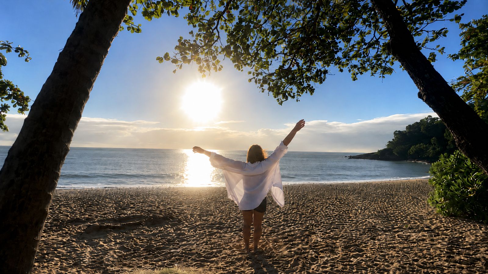 A woman with arms raised to the sunrise on a tropical beach, framed by trees
